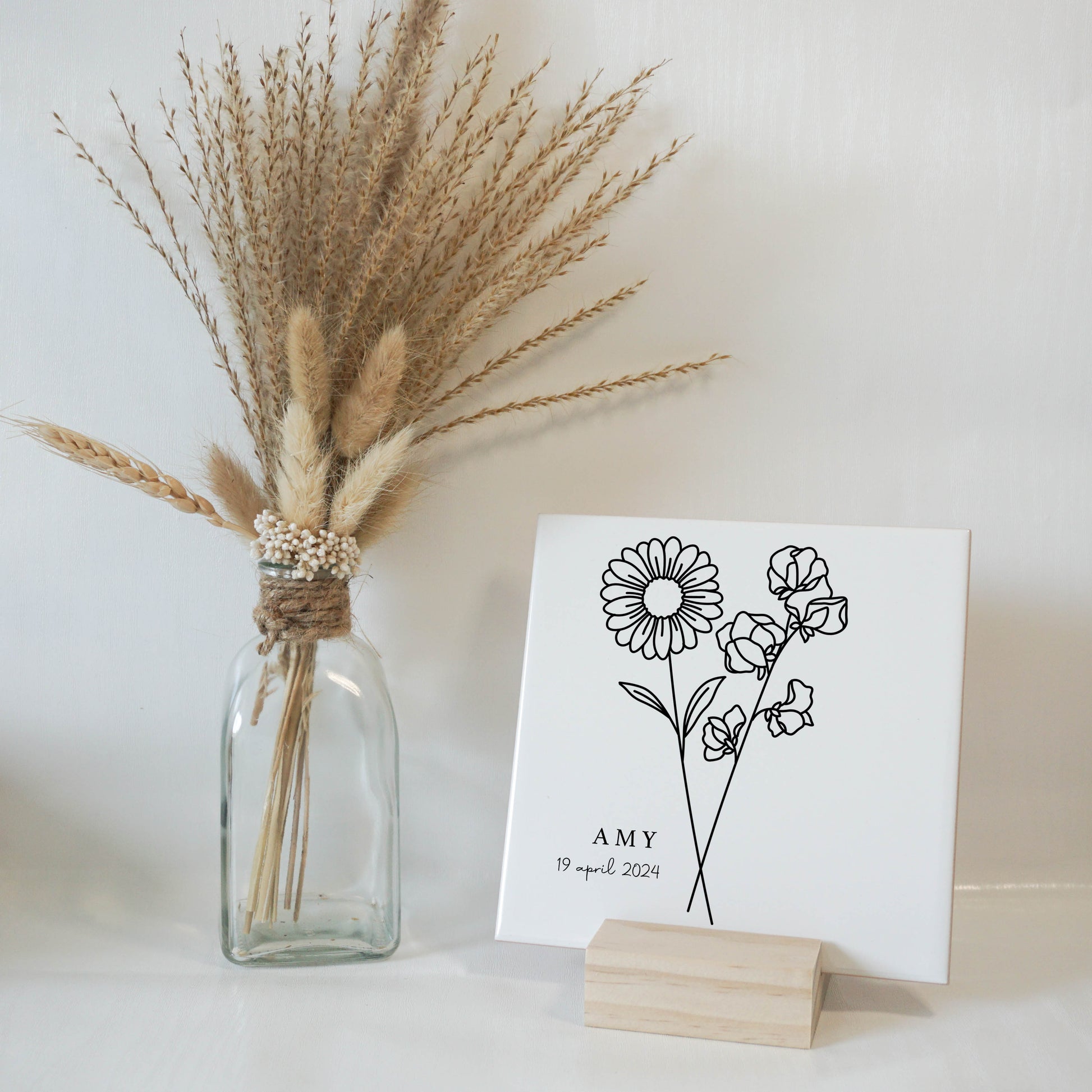 Close-up van een gepersonaliseerde keramische tegel met de april geboortebloemen (margriet en lathyrus), de naam [naam kind] en de geboortedatum.